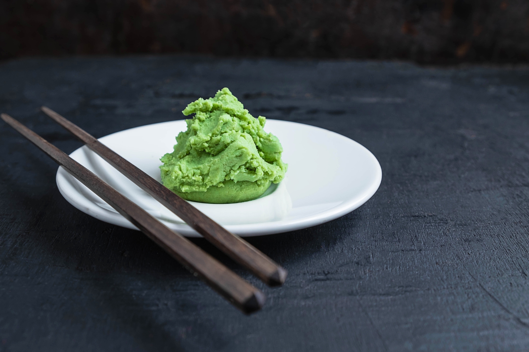 Bright green wasabi paste on a white plate with a pair of dark brown chopsticks beside.