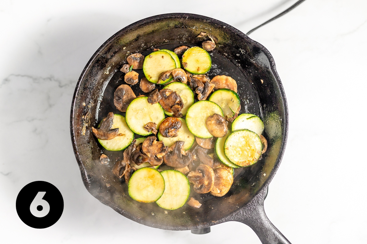 The zucchini and mushrooms in the black pan are softening and looking slightly darker in color.