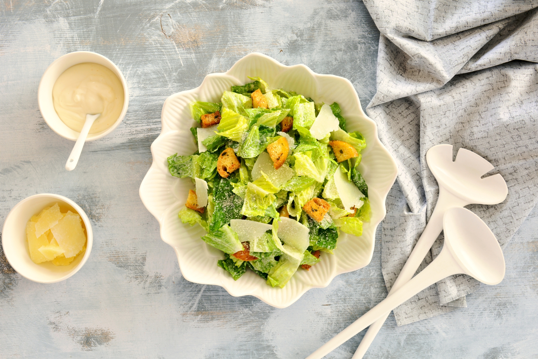 Scalloped edge white bowl of chopped romaine lettuce and croutons. There are white salad tongs, a bowl of creamy dressing and a bowl of parmesan cheese shards beside the bowl.