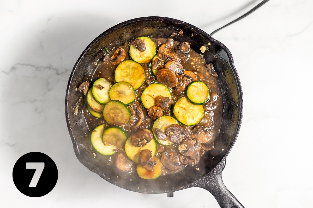 Soy sauce and other seasoning have been added to the cast iron pan of mushrooms and zucchini and the sauce looks very dark brown.