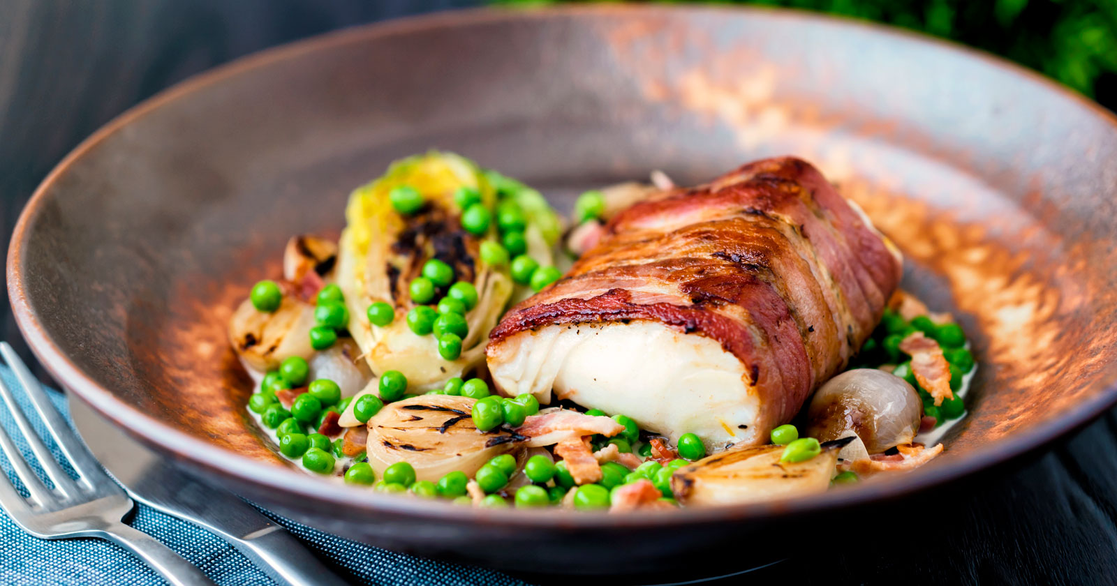 Frying pan with bacon wrapped white cod loin, peas and onions.