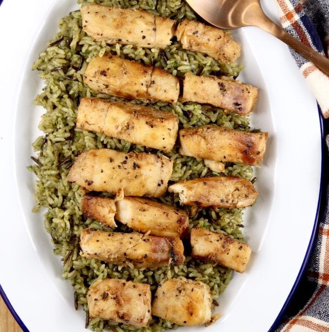 Rows of grilled cod on top of green seasoned rice on a large white oval platter.