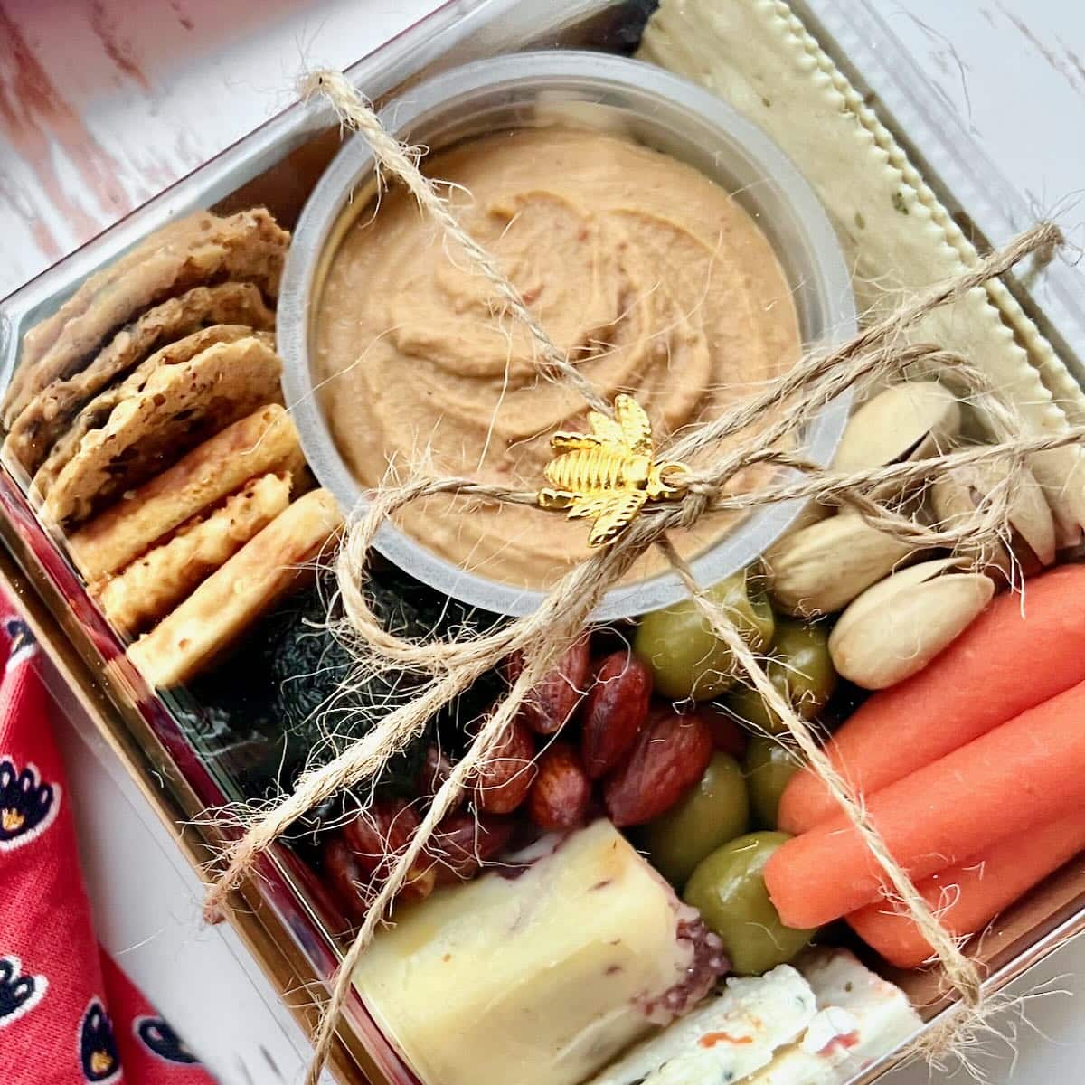 Open box of hummus, crackers, dates, pistachios, grapes, carrots and cheese tied up with jute twine.