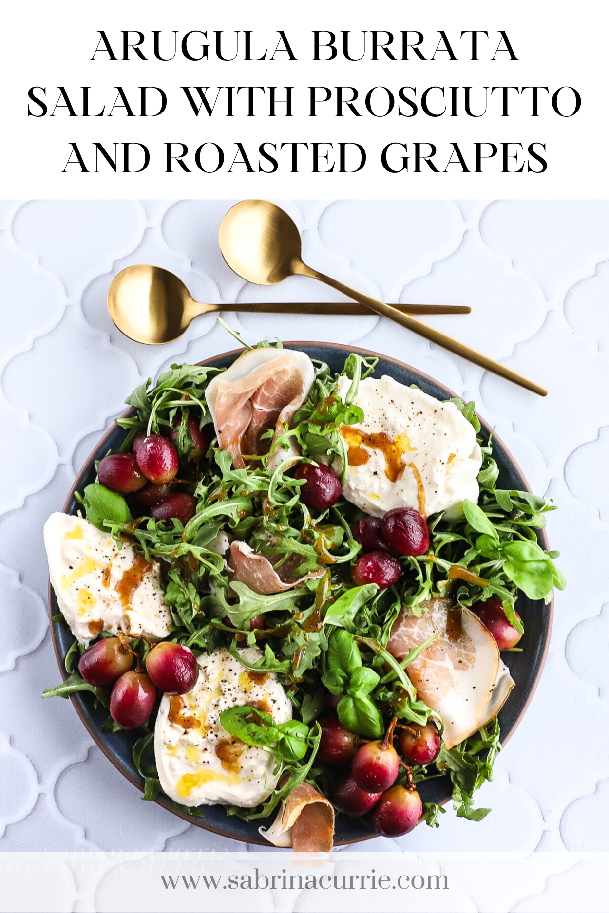 A round plate of green arugula, chunks of white burrata cheese, roasted purple grapes and prosciutto. Two gold spoons are above the plate. Recipe title is at the top.