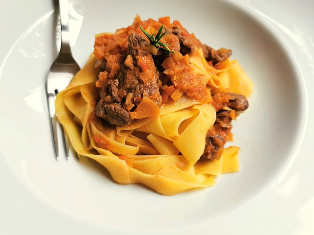 Round tangle of pappardelle noodles topped with red shredded boar ragu on a white plate. 