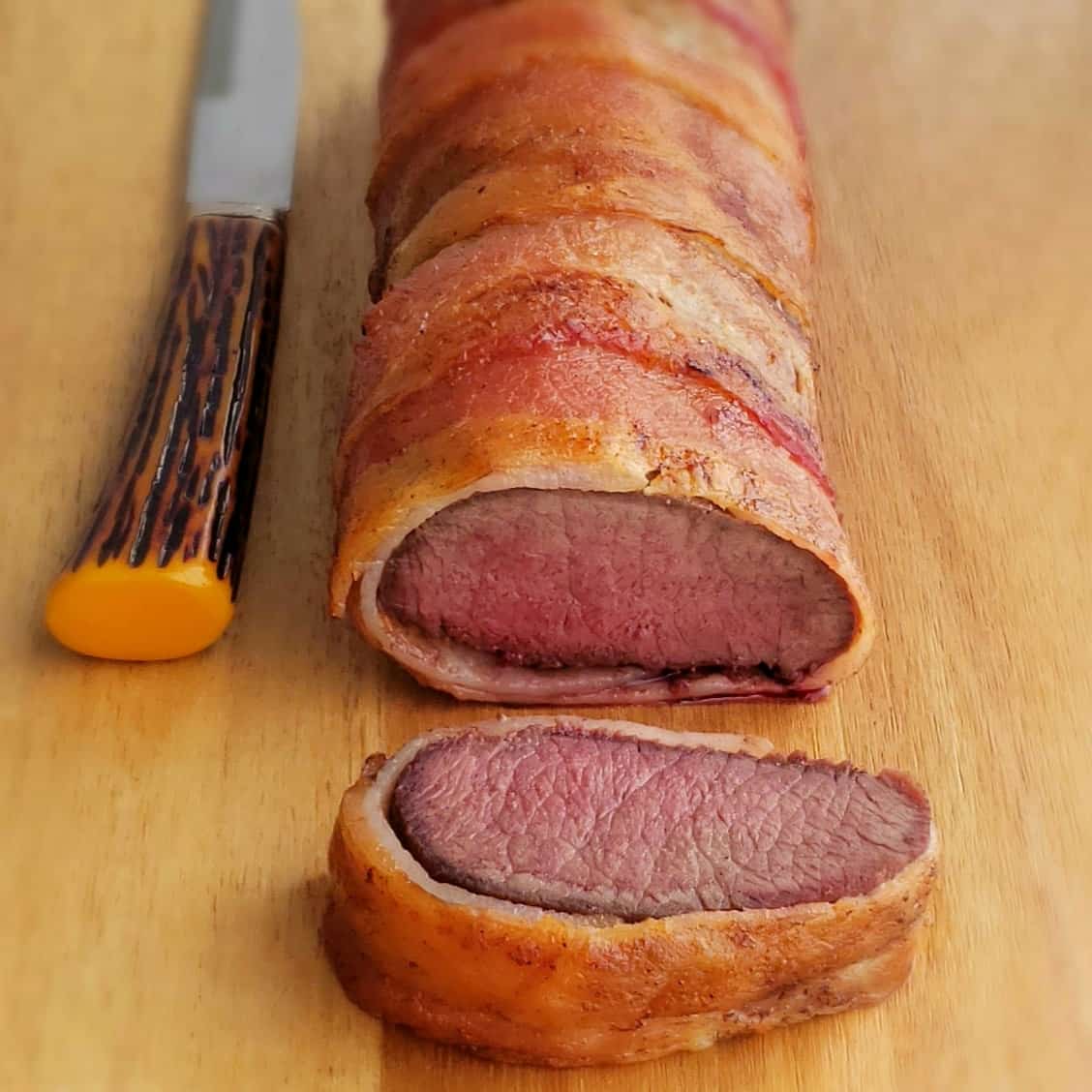 Bacon wrapped venison loin that is long and round with a knife beside. One round slice has been cut off the front showing the medium rare pink meat inside.