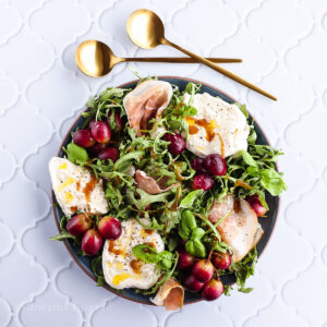 A round plate of green arugula, chunks of white burrata cheese, roasted purple grapes and prosciutto. Two gold spoons are above the plate and some torn chunks of bread are beside the plate.