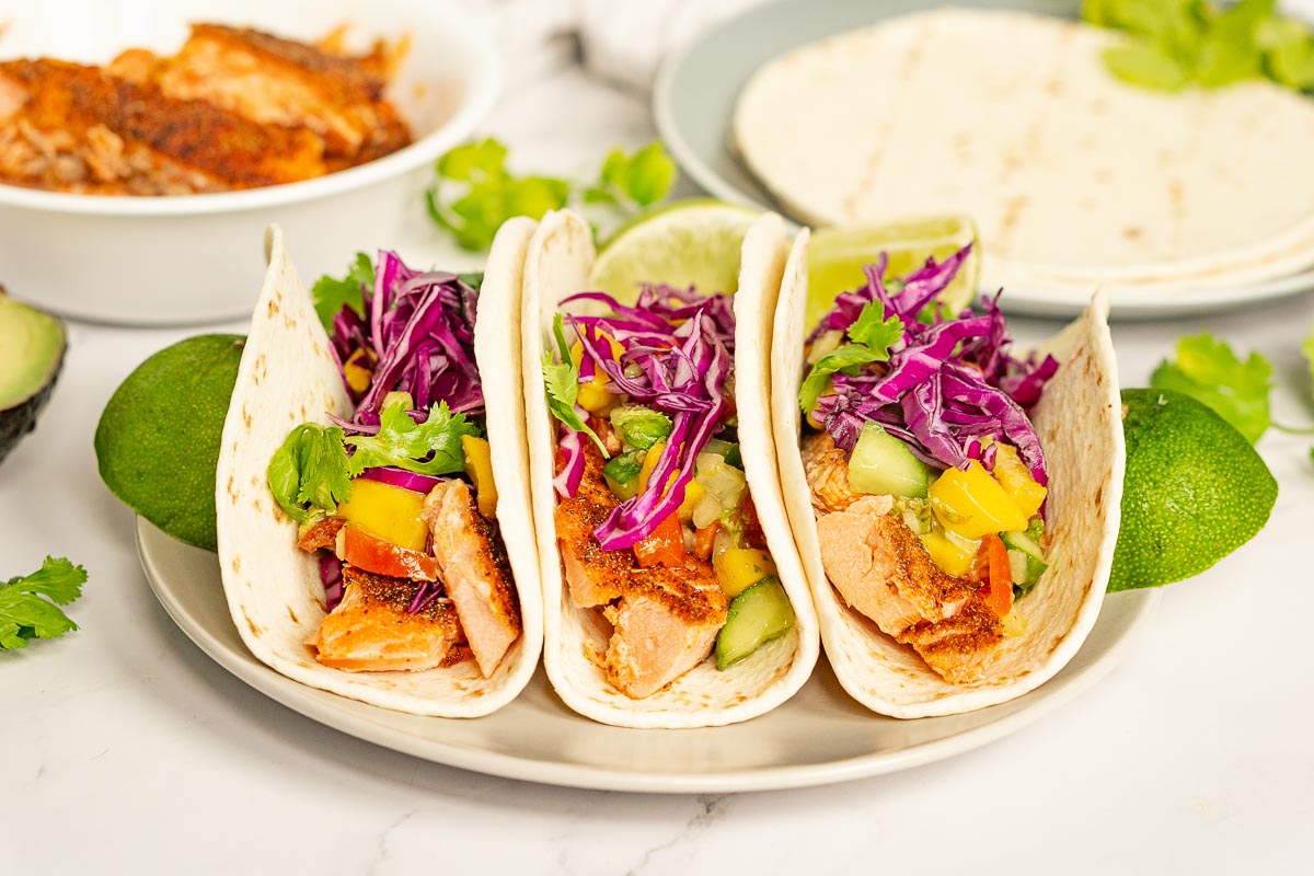 3 White flour tortilla tacos filled with colorful cabbage slaw, fried fish and yellow fruit.