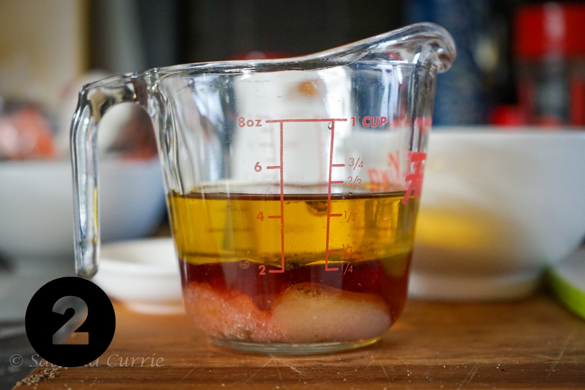 A glass measuring cup with a layer of salt, pepper, mustard and sugar, a layer of red wine vinegar and the top layer is clear, yellow orange olive oil.