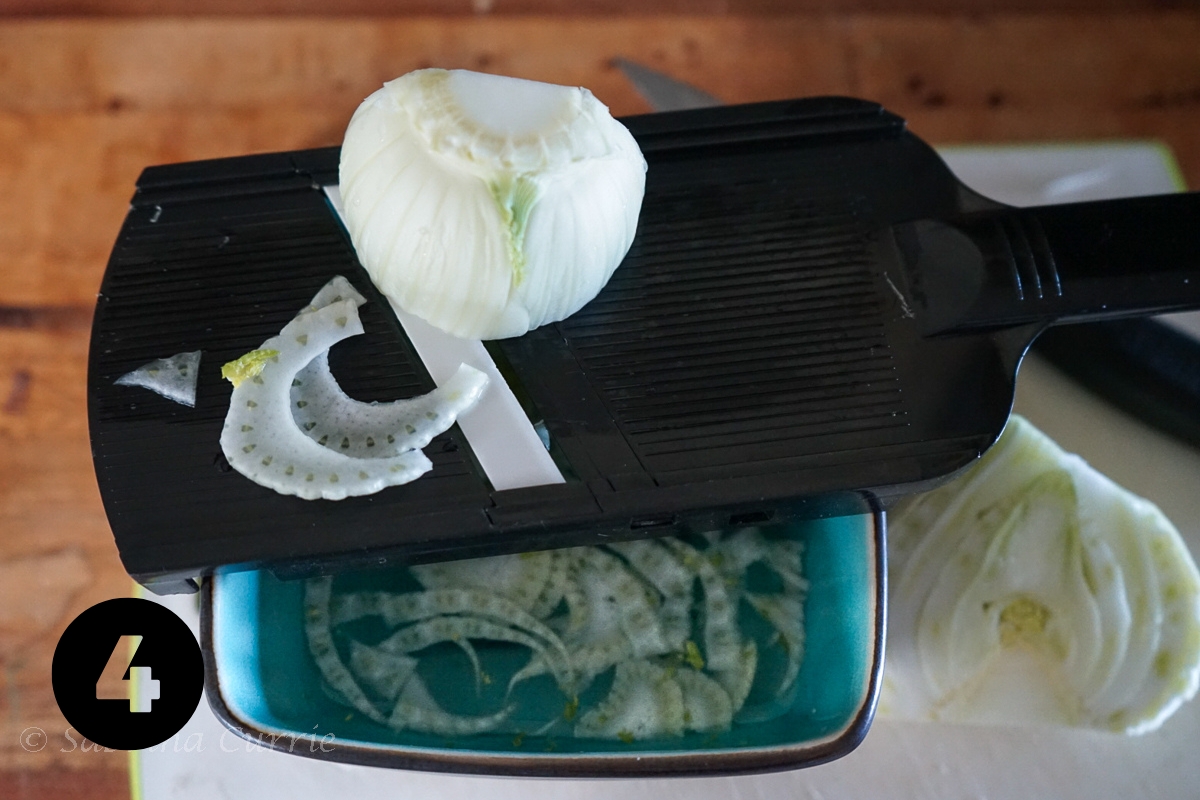 A trimmed bulb of pale green and white fennel on a black mandolin with thin slices of fennel in a blue bowl below.