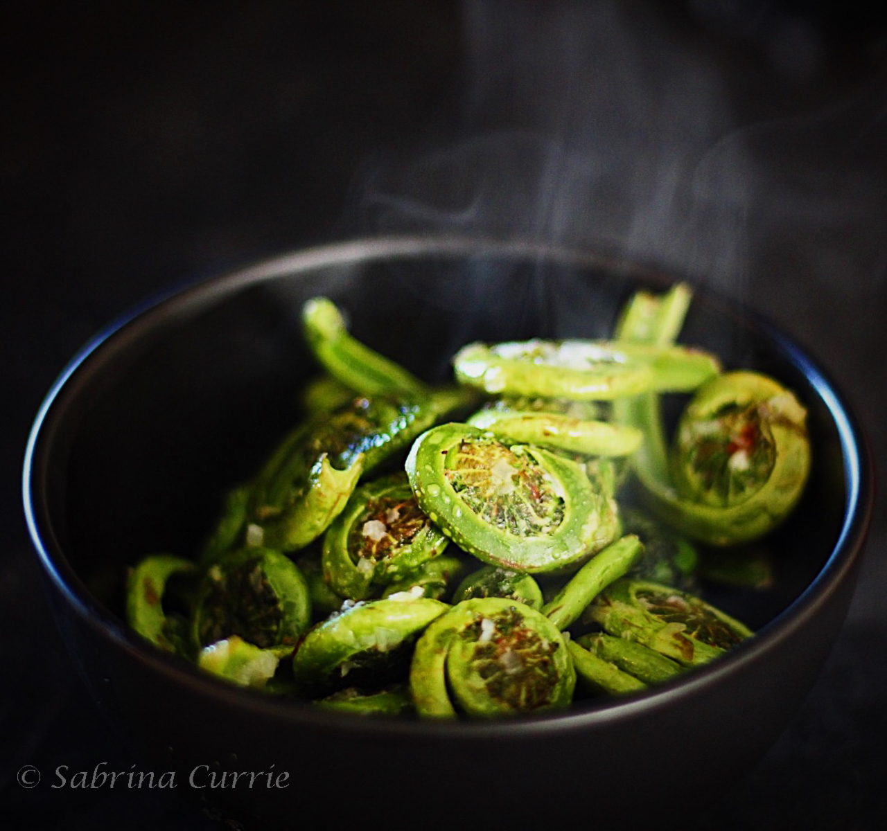 Black bowl of bright green fiddleheads cooked and steaming with flecks of chili and garlic on top.