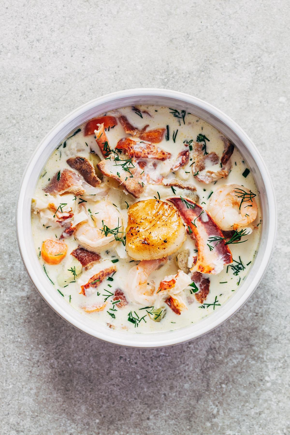 Creamy white soup in a white bowl full of bright orange, pink and golden brown chunks of seafood including lobster, scallops and white fish.