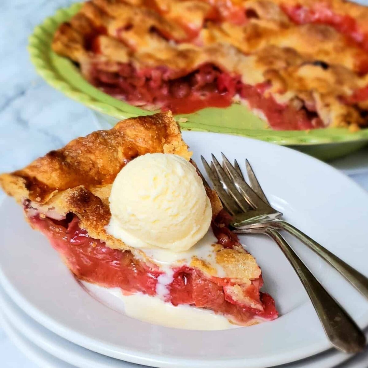 Slice of pie with pink strawberry rhubarb filling and classic pastry crust topped with a round scoop of vanilla ice cream.