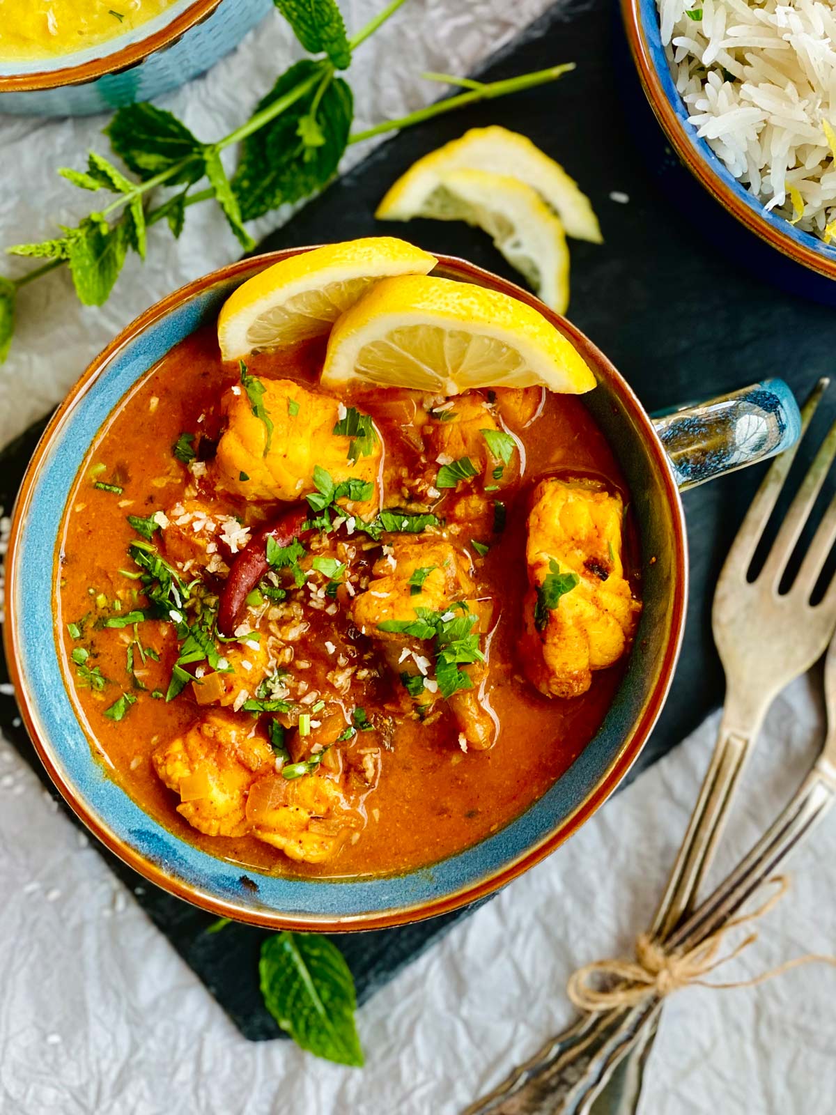 Birdsview Monkfish curry garnished with two slices of fresh lemon and fresh herbs served in a blue soup bowl.