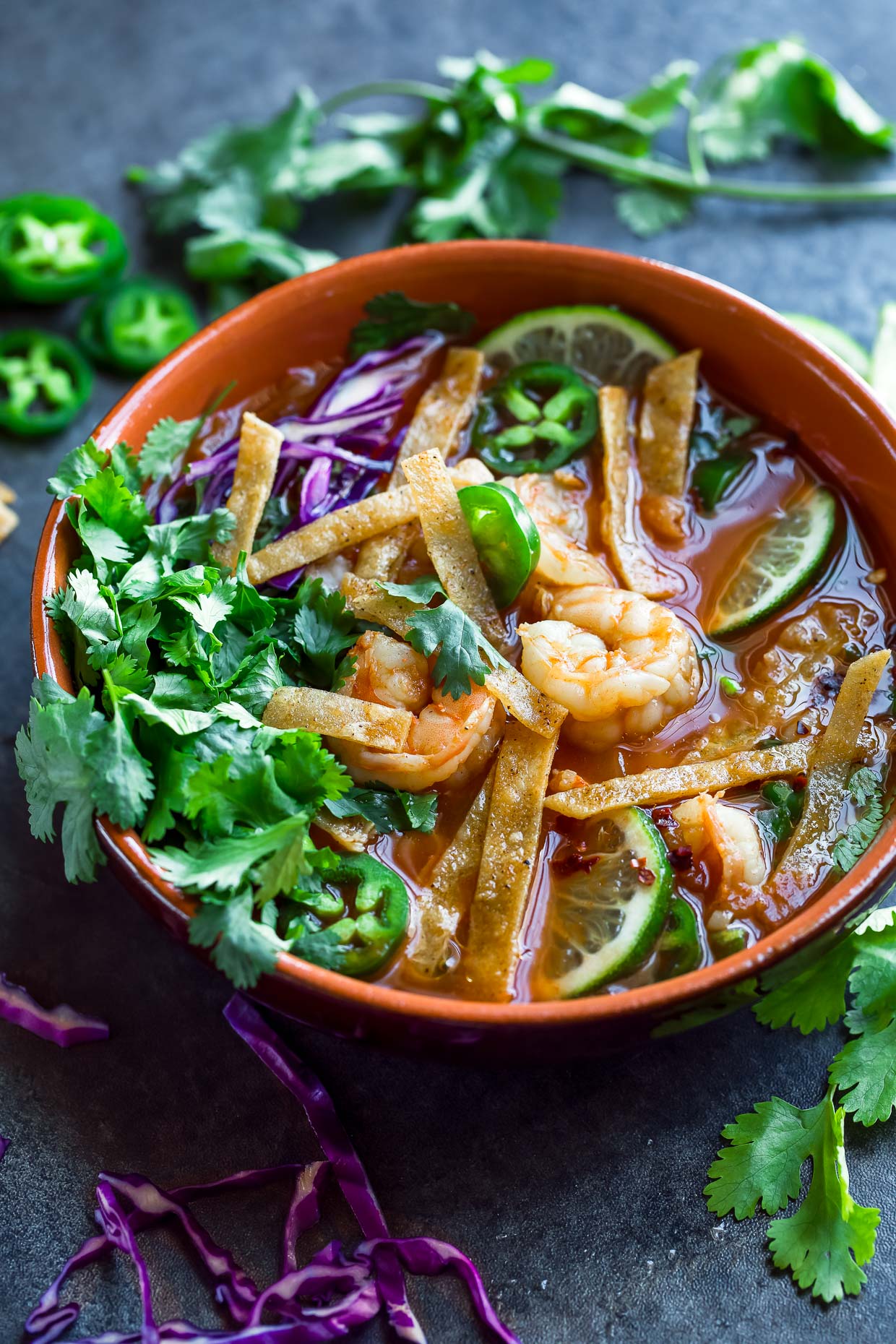 Orange terracotta bowl of shrimp and tortilla soup topped with shredded purple cabbage and chopped green cilantro.