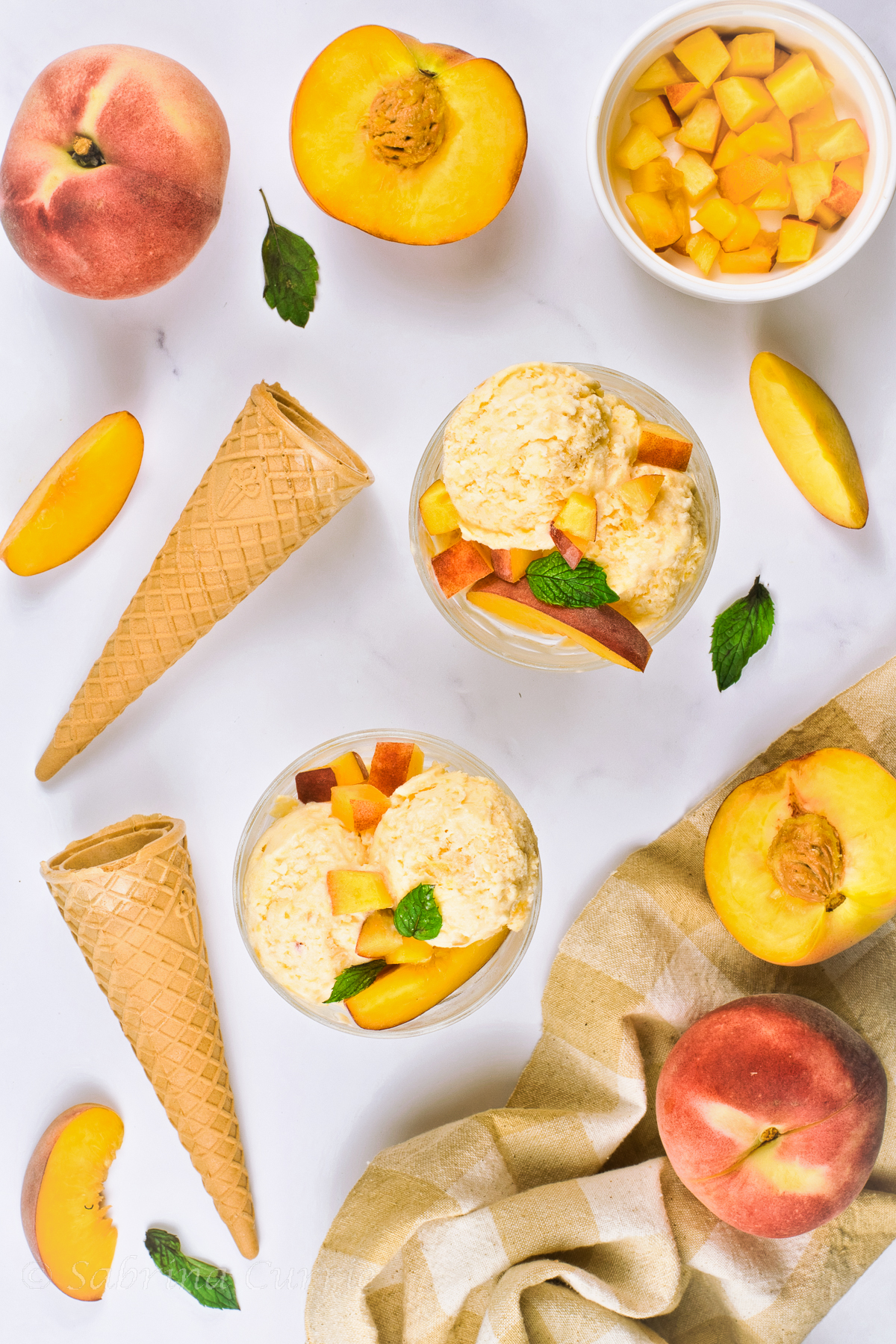 Waffle cones, whole and chopped peaches and mint leaves scattered around 2 glass bowls of pale yellow ice cream.