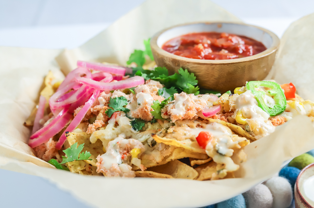 Cheesy nachos topped with chunks of lobster, red pepper and pickled onions with a bowl of salsa on one side.
