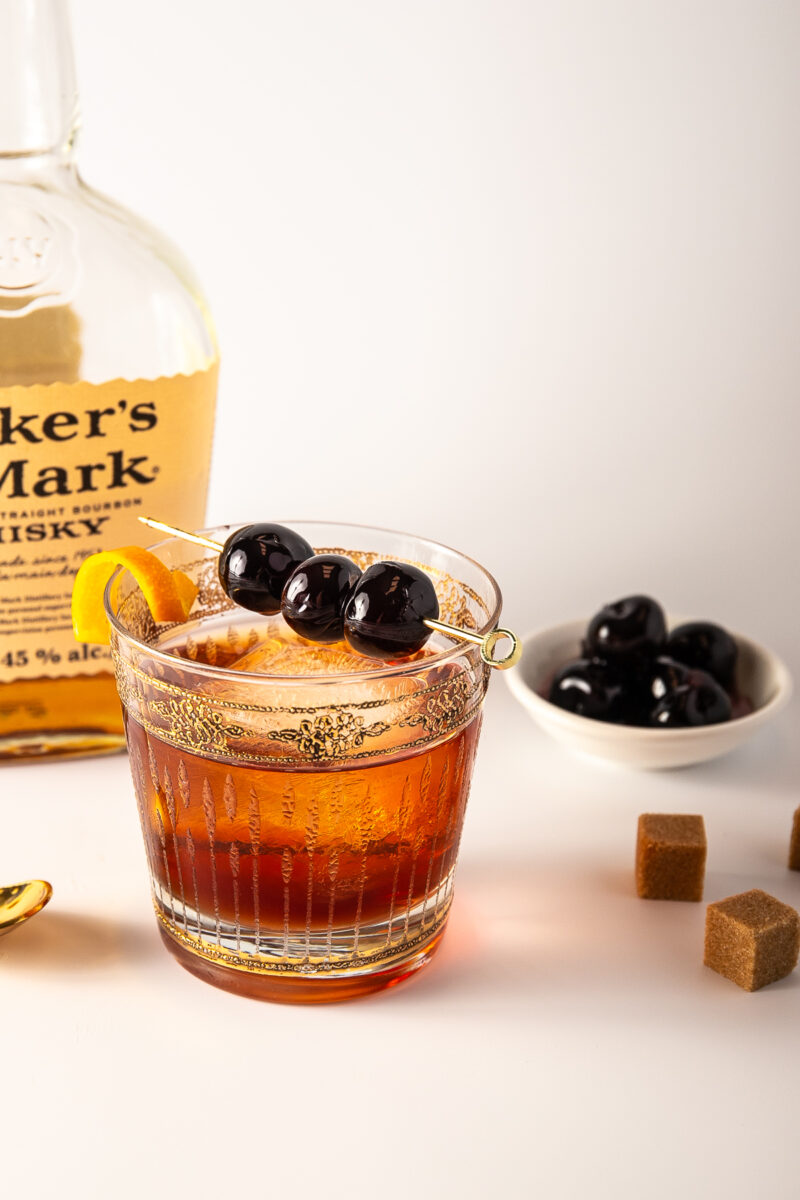 A cocktail topped with cherries and orange peel in front of a bottle of Maker's Mark bourbon and a dish of extra cherries.