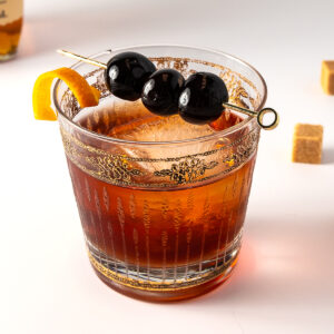 A rocks glass with one large ice cube and a dark drink in it with a cocktail pick of cherries on top and a twist of orange on the rim.