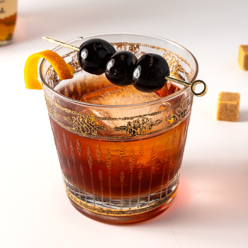A rocks glass with one large ice cube and a dark drink in it with a cocktail pick of cherries on top and a twist of orange on the rim.