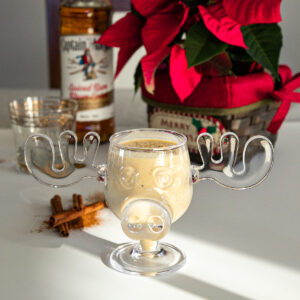 A moose mug of creamy eggnog in front of a Christmas flower arrangement and a bottle of spiced rum.