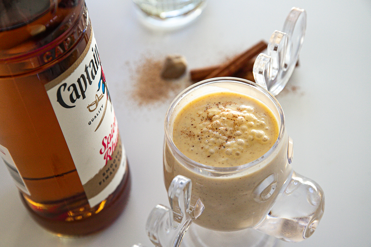 A clear moose mug full of frothy pale yellow eggnog and a bottle of spiced rum.