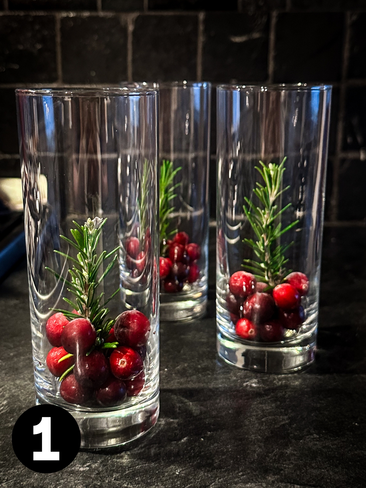 3 tall narrow glasses with cranberries in the bottom holding a sprig of rosemary upright in the middle.