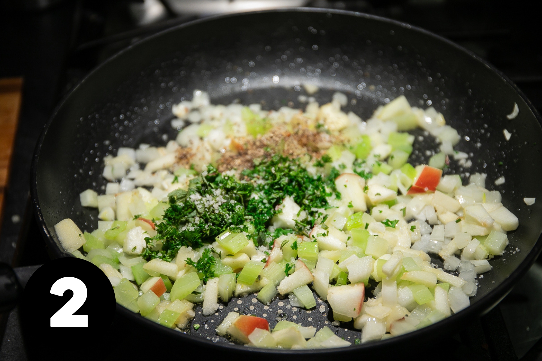 A frying pan of diced onion, celery and apple with chopped green herbs on top.