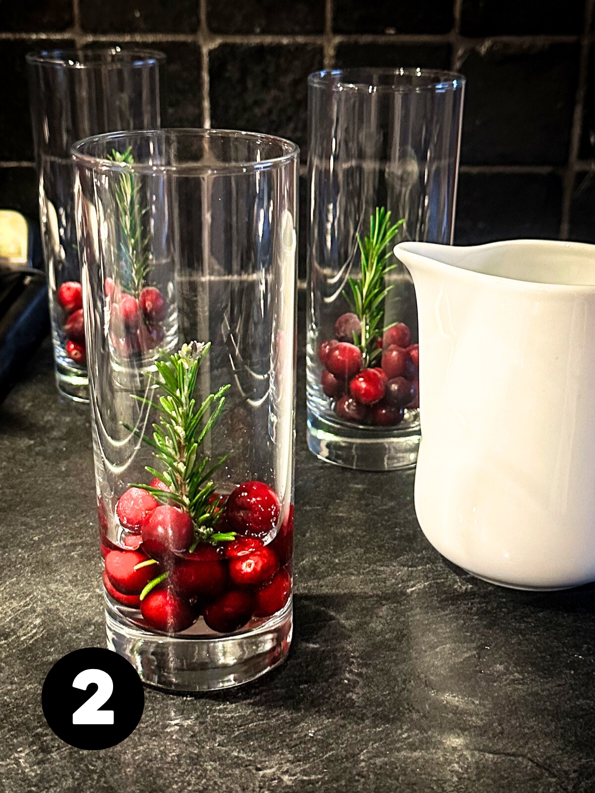 3 tall narrow glasses with cranberries in the bottom holding a sprig of rosemary upright in the middle and water coming up partway to cover the cranberries.