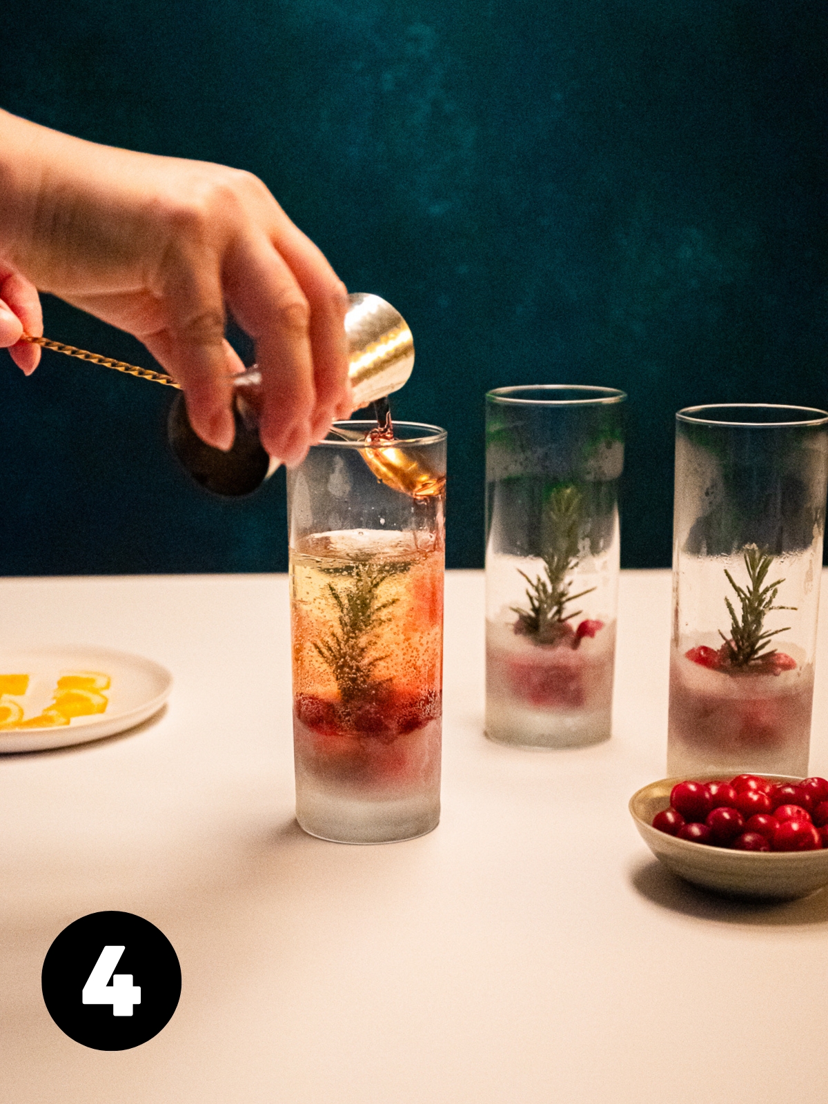 Cranberry juice being poured over the back of a bar spoon into a glass of prosecco with frozen cranberries and herb sprig in the bottom.