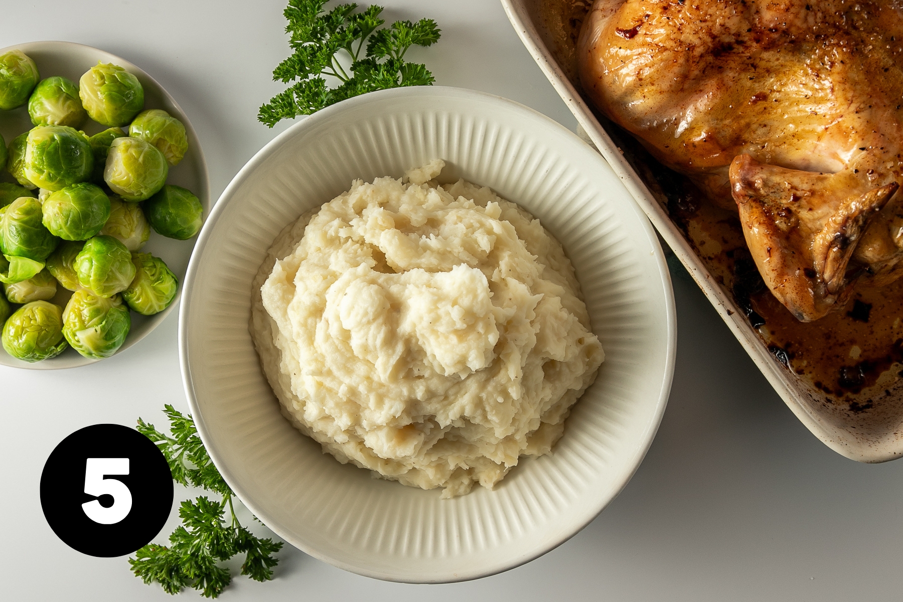 Mashed potatoes in a bowl alongside a roast chicken and steamed Brussels sprouts.