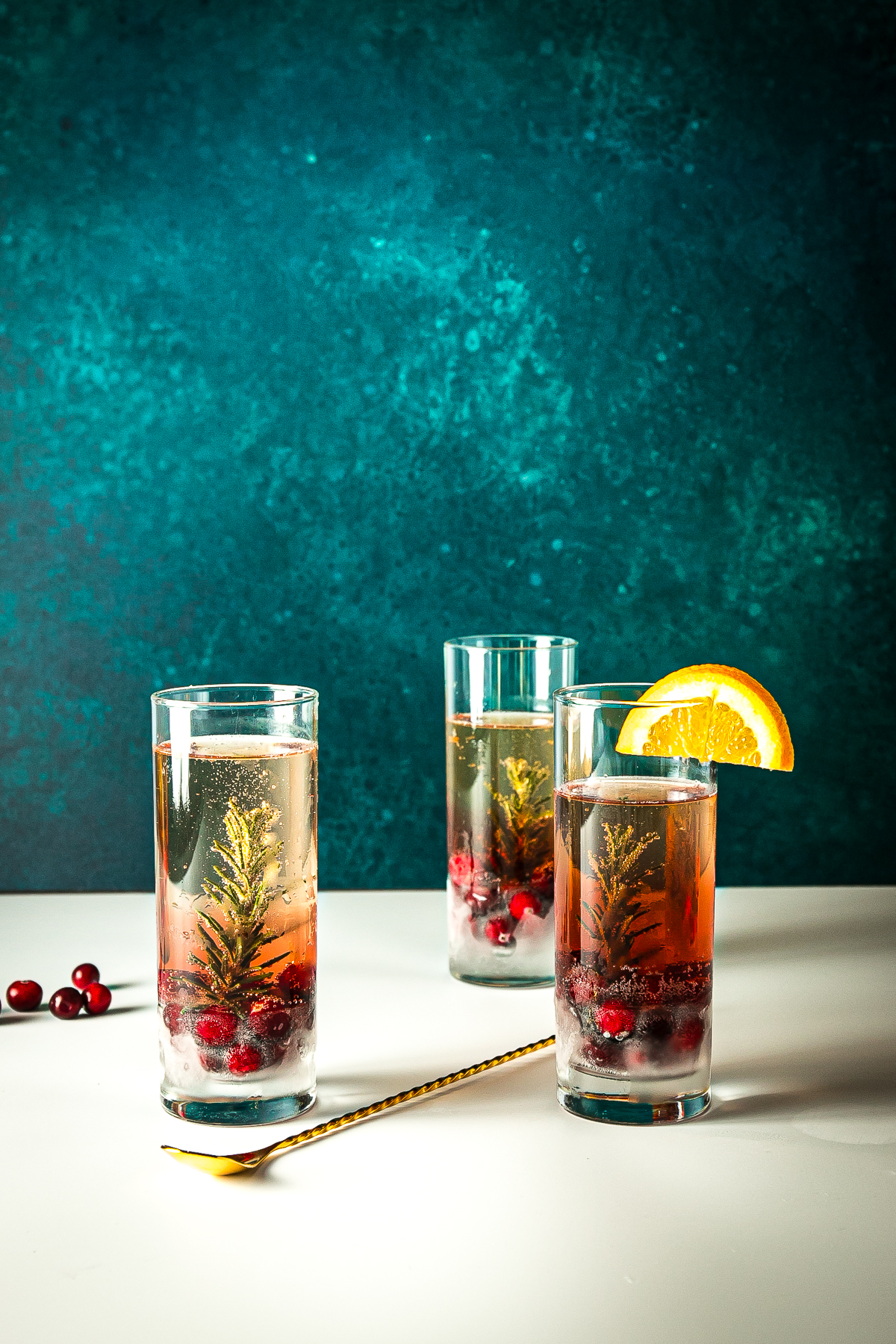 3 tall glasses each with a sprig of rosemary, cranberries and effervescent prosecco in them and a half wheel of orange on one of them.