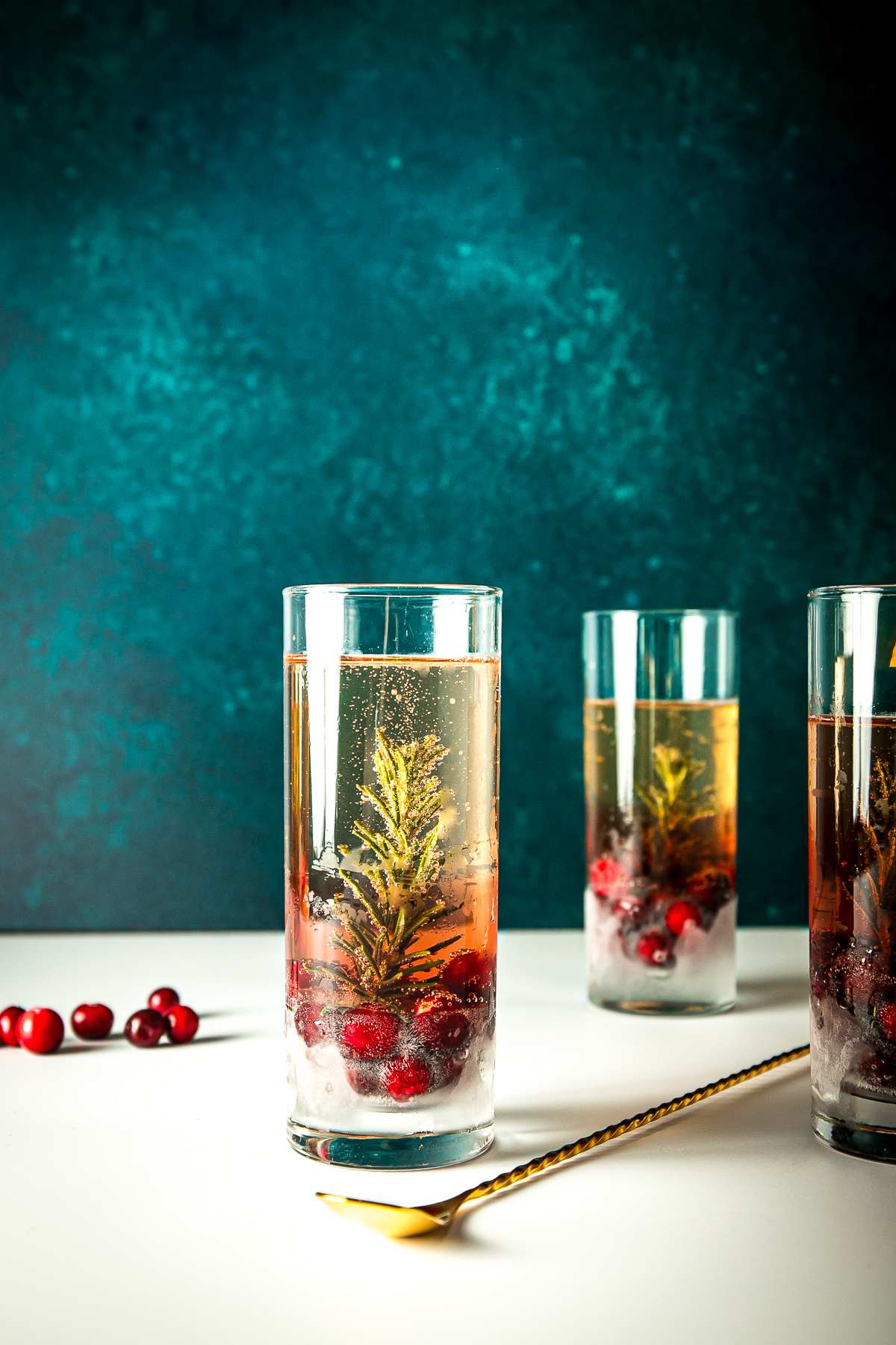 3 tall glasses each with a sprig of rosemary, cranberries and effervescent prosecco in them.