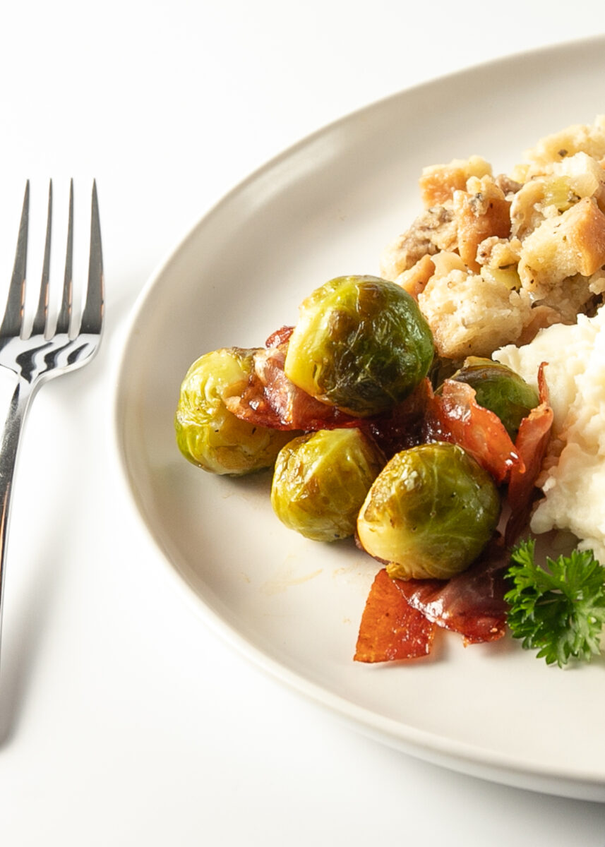 5 cooked Brussels sprouts with shards of crispy prosciutto on a plate with bread stuffing and mashed potatoes.