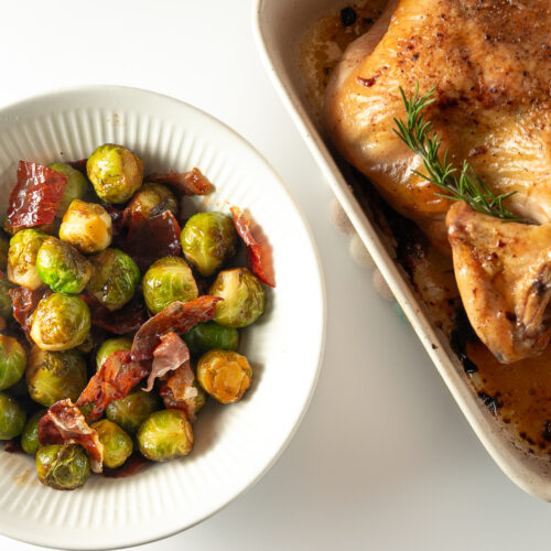 A bowl of cooked Brussels sprouts and prosciutto beside a roast chicken in a pottery roasting pan.