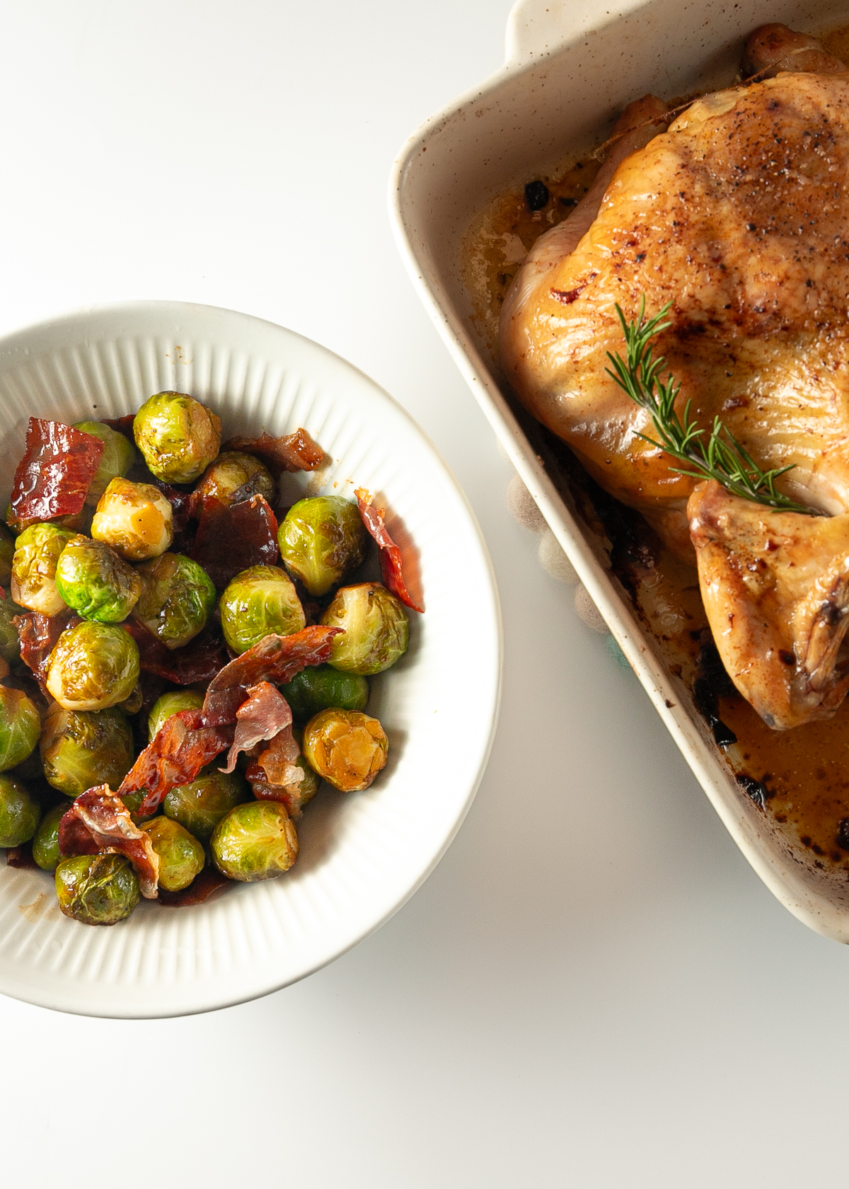 A bowl of cooked Brussels sprouts and prosciutto beside a roast chicken in a pottery roasting pan.