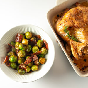 A bowl of roasted Brussels sprouts and prosciutto beside a roast chicken in a pottery roasting pan.