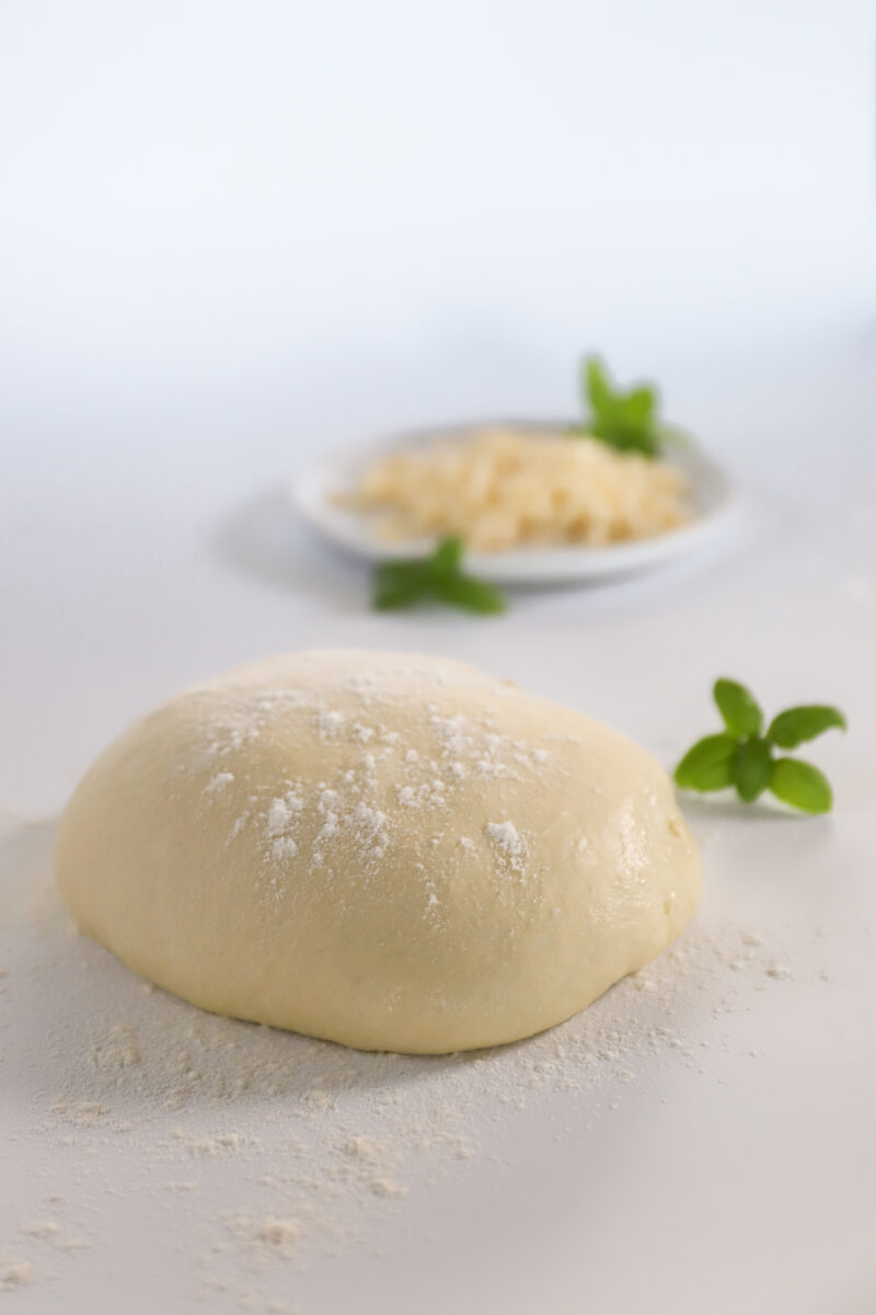 A dough ball dusted with flour and a bowl of shredded mozzarella cheese with a few basil leaves scattered around.