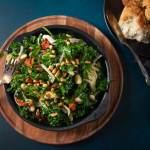 Bowl of salad on a wooden charger with a plate of bread beside.