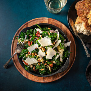A chopped kale salad with white beans, shards of parmesan cheese, toasted pine nuts, slivered red onion and sundried tomatoes in a bowl beside some torn crusty bread and a glass of white wine.