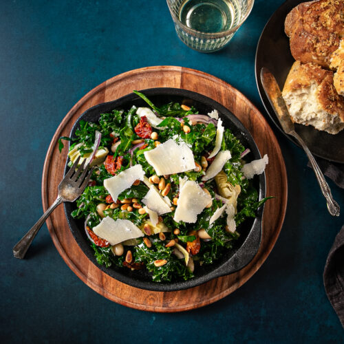 A chopped kale salad with white beansshards of parmesan cheesetoasted pine nutsslivered red onion and sundried tomatoes in a bowl beside some torn crusty bread and a glass of white wine.