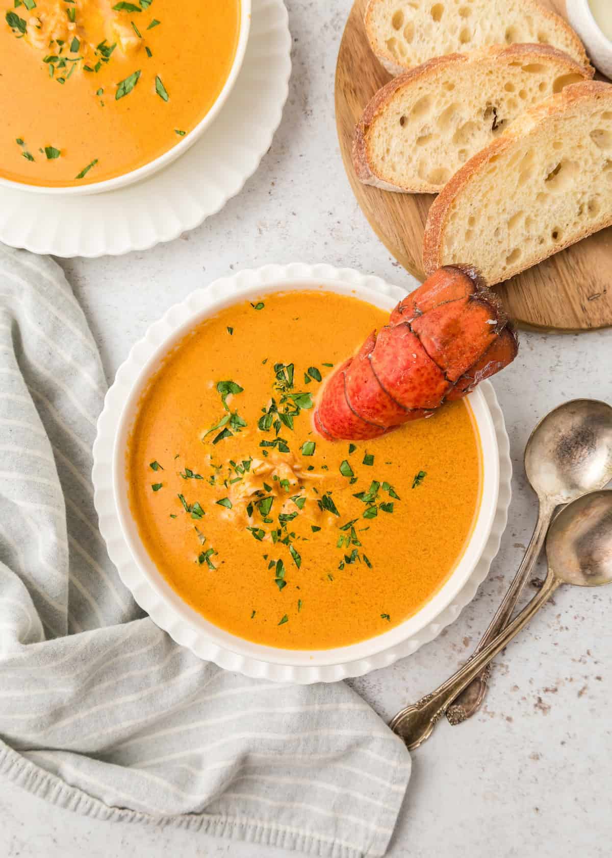 Bowls of lobster bisque garnished with chopped parsley and a little lobster meat. One bowl of soup has a lobster tail sticking out of it and there is a plate of bread beside.