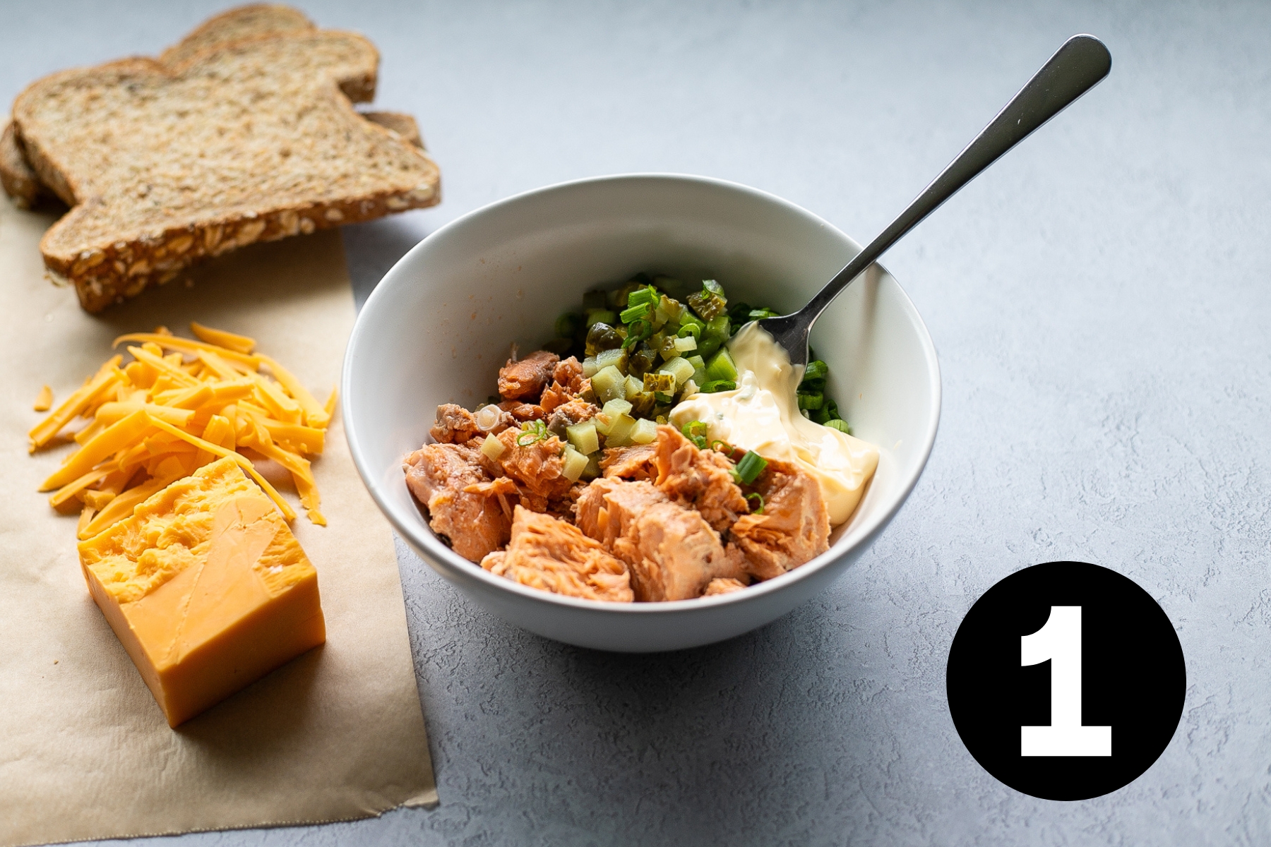 Salmon, mayonnaise and chopped veggies in a bowl beside 2 slices of brown bread and some shredded cheddar cheese.