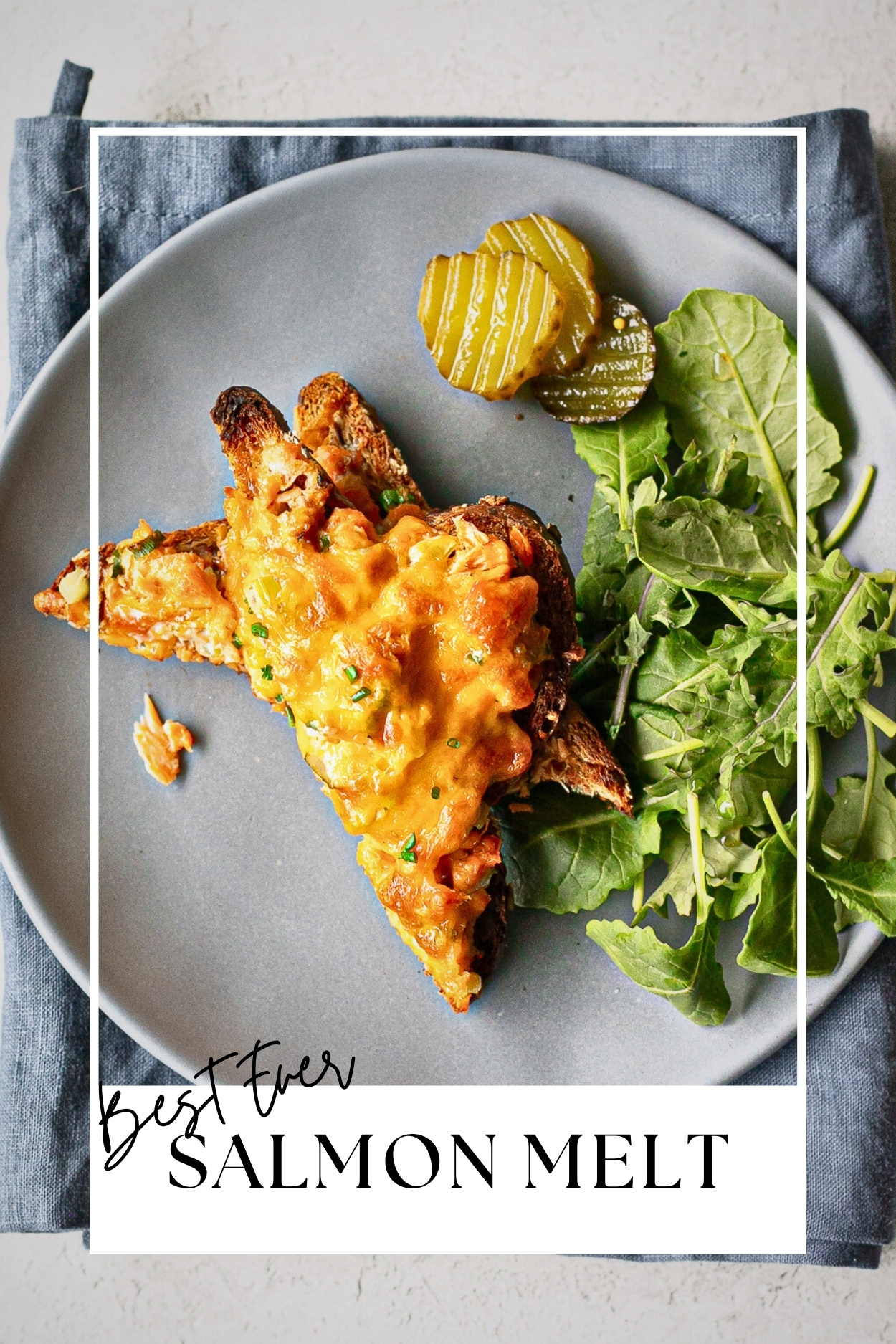 Plated halves of a salmon melt with a tossed green salad and pickle slices.