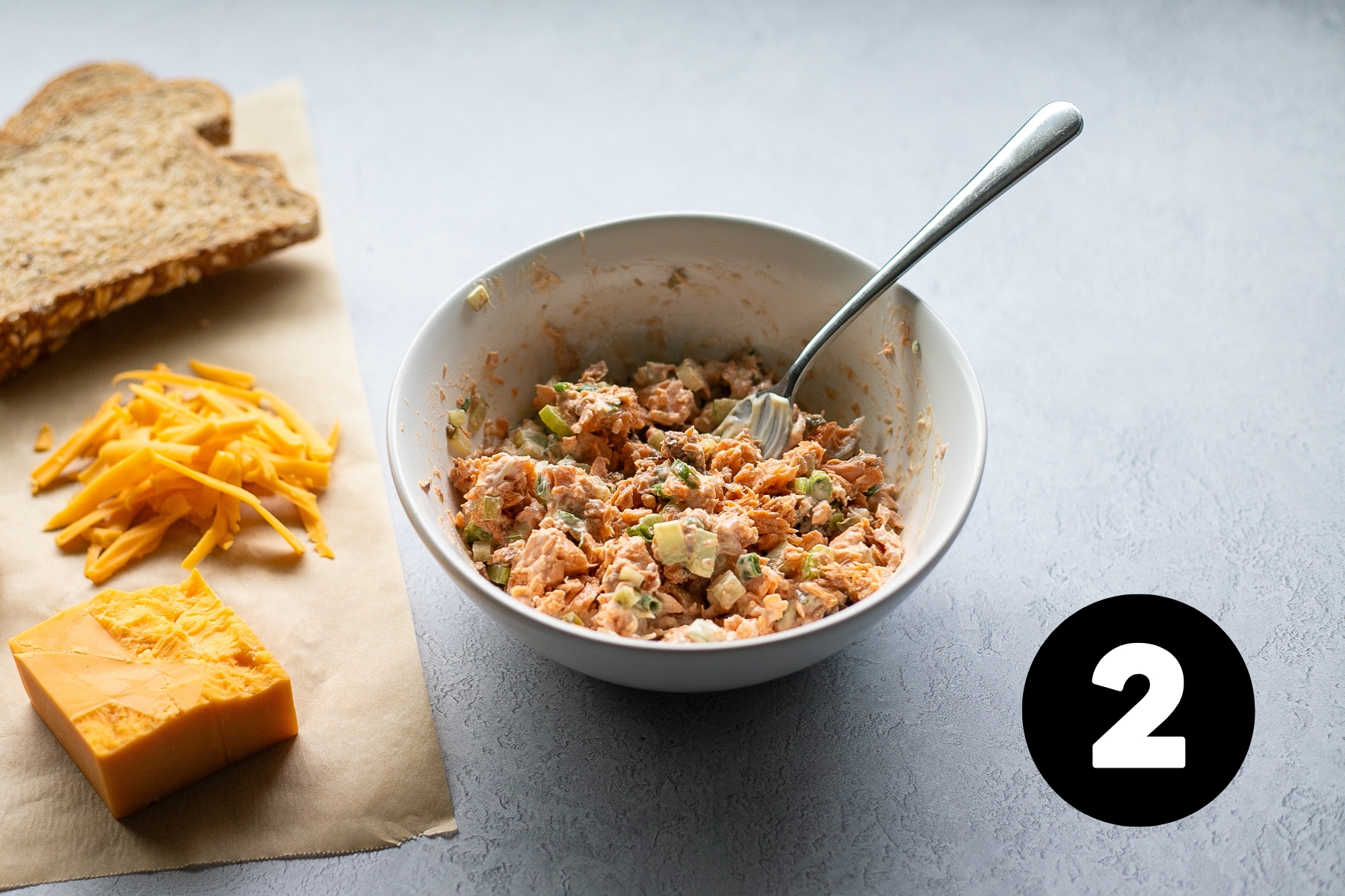 Mixed salmon salad in a bowl beside 2 slices of brown bread and some shredded cheddar cheese.