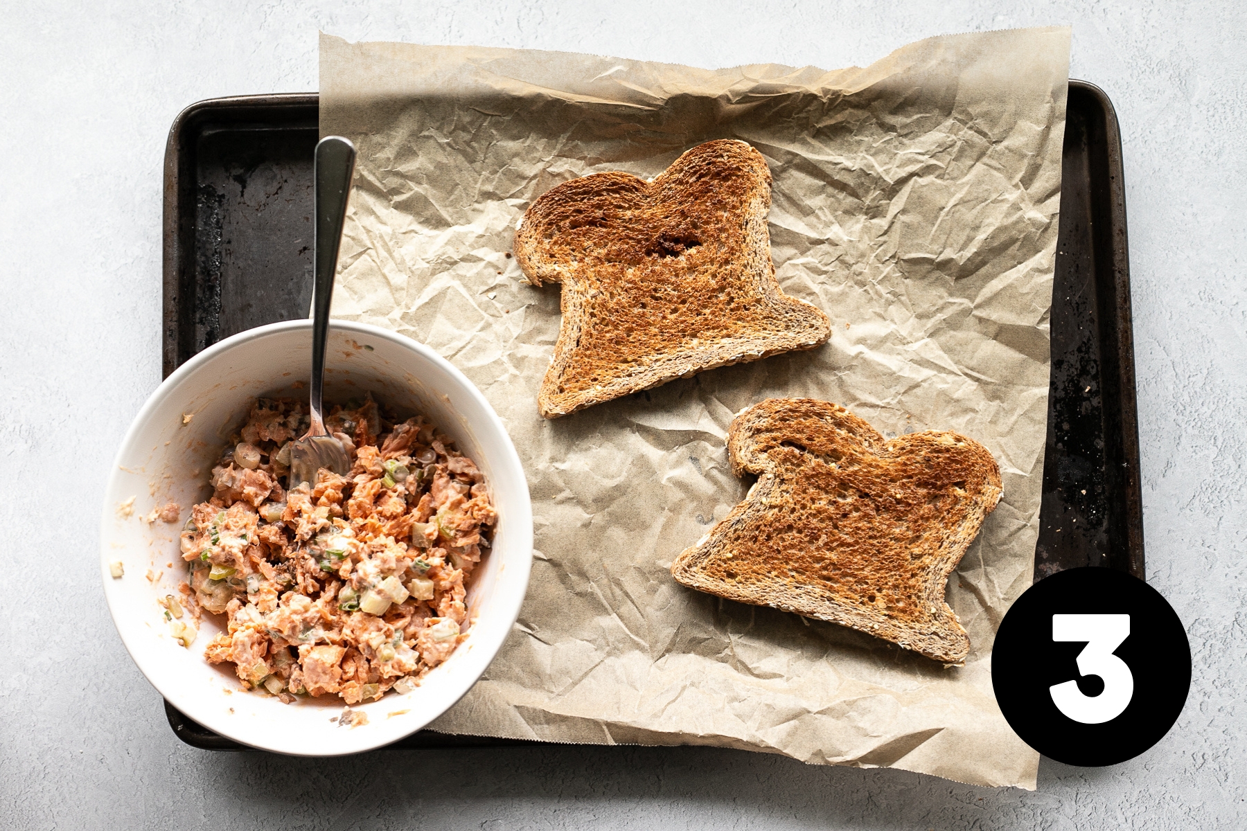 Two pieces of toasted bread on a parchment lined baking sheet with the salmon mixture in a bowl beside.