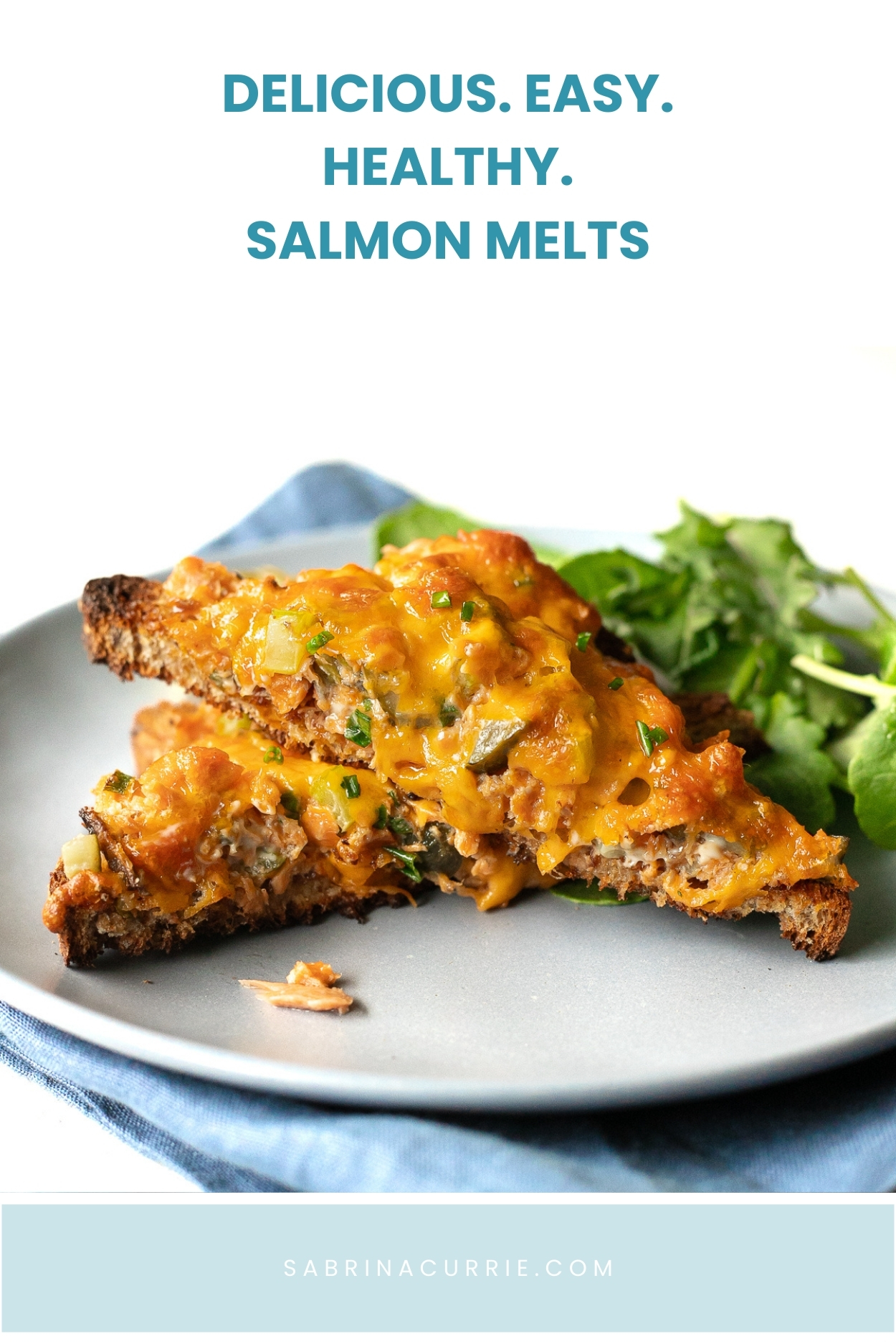 A salmon melt toast cut in half into 2 triangles sitting on a plate with some green salad beside. The recipe title is above the plated sandwich.