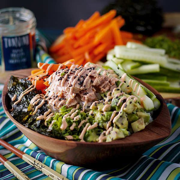 A bowl heaped with chopped vegetables and tuna and drizzled crosswise with a creamy sauce.