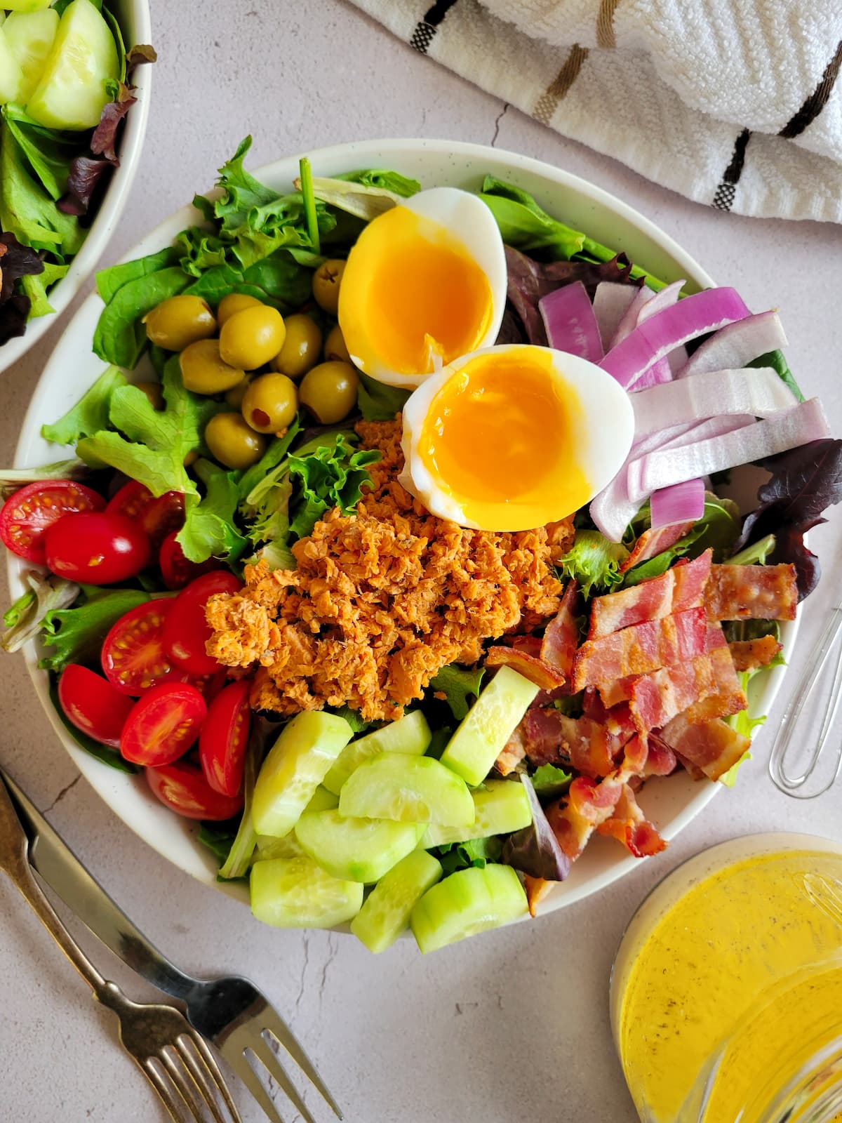 Composed salad in a bowl including a halved soft boiled egg, chopped vegetables, green olives, red onion and salmon salad.