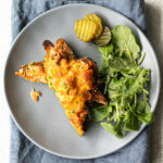 Green salad, sliced pickles and two triangular halves of a salmon melt on a plate that is placed on a cloth napkin.
